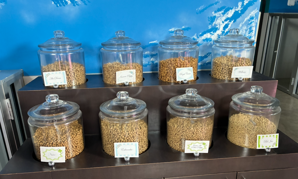 Row of glass jars with lids on dark wooden shelves, filled with cereals and nuts, each with a small label plate in front
